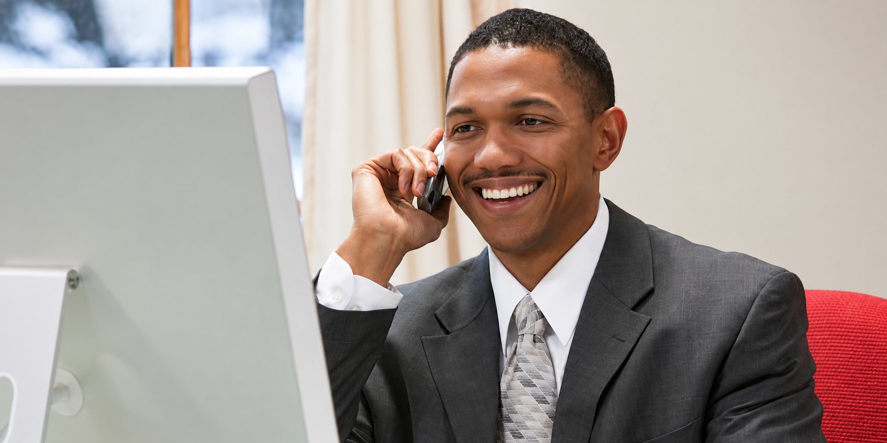 Un homme souriant en costume gris et cravate parle au téléphone, assis à un bureau devant un écran d’ordinateur.